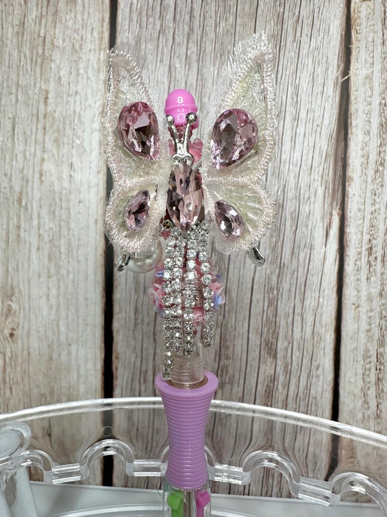 Puede incluir: Un bol&iacute;grafo decorativo con dise&ntilde;o de mariposa. La mariposa tiene alas de encaje blanco adornadas con gemas rosas. El cuerpo del bol&iacute;grafo es lavanda y el cuerpo de la mariposa est&aacute; adornado con pedrer&iacute;a.
