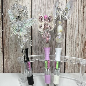 Puede incluir: Tres bol&iacute;grafos decorativos con adornos de mariposa en un soporte de pl&aacute;stico transparente. Los bol&iacute;grafos tienen detalles en negro, lavanda y blanco. Los adornos de mariposa est&aacute;n adornados con cristales y cuentas. Los bol&iacute;grafos se exhiben sobre un fondo de madera.