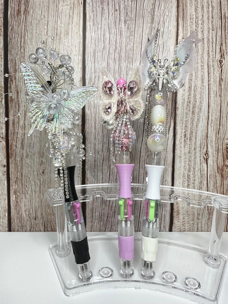 Puede incluir: Tres bol&iacute;grafos decorativos con adornos de mariposa en un soporte de pl&aacute;stico transparente. Los bol&iacute;grafos tienen detalles en negro, lavanda y blanco. Los adornos de mariposa est&aacute;n adornados con cristales y cuentas. Los bol&iacute;grafos se exhiben sobre un fondo de madera.
