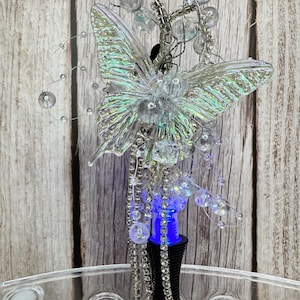 Puede incluir: Un tap&oacute;n de botella decorativo con un dise&ntilde;o de mariposa transparente e iridiscente. La mariposa est&aacute; adornada con cuentas transparentes y detalles de alambre plateado. El tap&oacute;n tiene una base negra con luz azul y un interior rosa.