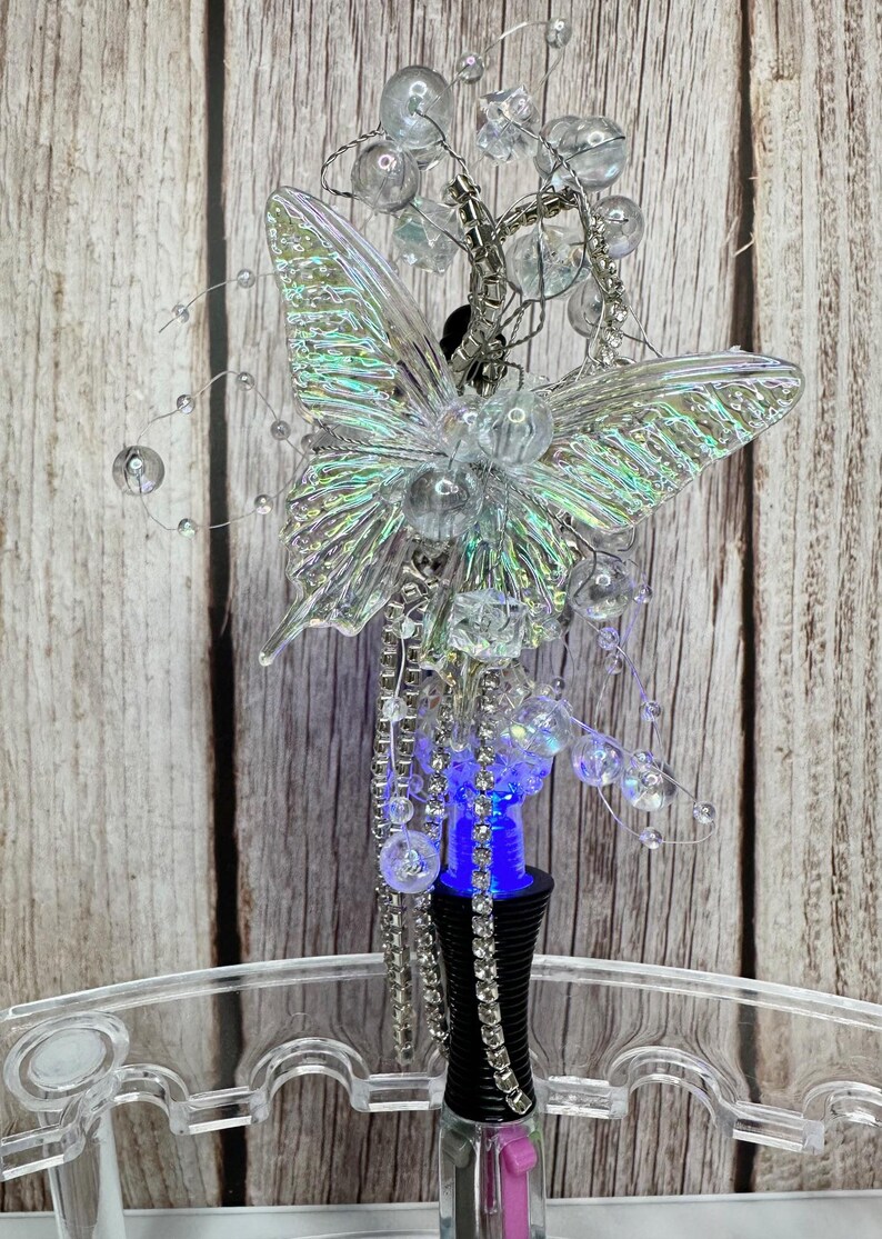 Puede incluir: Un tap&oacute;n de botella decorativo con un dise&ntilde;o de mariposa transparente e iridiscente. La mariposa est&aacute; adornada con cuentas transparentes y detalles de alambre plateado. El tap&oacute;n tiene una base negra con luz azul y un interior rosa.