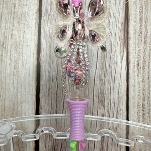 Puede incluir: Bol&iacute;grafo decorativo con dise&ntilde;o de mariposa. La mariposa tiene detalles de cristal rosa y transparente, con cabeza y alas rosas. El cuerpo del bol&iacute;grafo es lavanda y contiene varios colores de tinta.