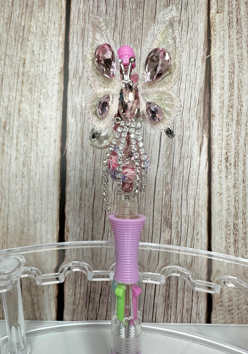 Puede incluir: Bol&iacute;grafo decorativo con dise&ntilde;o de mariposa. La mariposa tiene detalles de cristal rosa y transparente, con cabeza y alas rosas. El cuerpo del bol&iacute;grafo es lavanda y contiene varios colores de tinta.