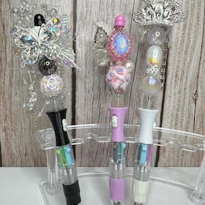 Puede incluir: Tres bol&iacute;grafos decorativos con adornos ornamentados. Un bol&iacute;grafo tiene un dise&ntilde;o de mariposa, otro una joya rosa y el tercero una corona plateada. Los bol&iacute;grafos tienen barriles negros, lavanda y blancos, y se exhiben en un soporte de pl&aacute;stico transparente.