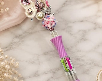 Op de afbeelding: Een decoratieve pen met een vlinder- en bloemversiering. De pen heeft een transparante schacht met roze, groene en magenta inktpatronen. De vlinder is versierd met roze edelstenen en parels. De pen is felroze.