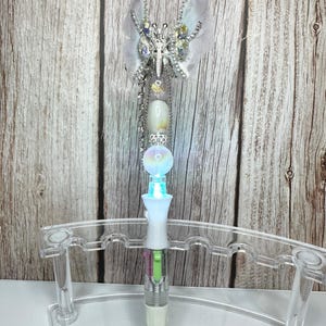 Puede incluir: Un bol&iacute;grafo decorativo con dise&ntilde;o de mariposa. El bol&iacute;grafo presenta una mariposa plateada con detalles transparentes e iridiscentes. El cuerpo del bol&iacute;grafo tiene un dise&ntilde;o blanco, transparente e iridiscente con una funci&oacute;n de iluminaci&oacute;n. El bol&iacute;grafo se exhibe en un soporte de pl&aacute;stico transparente.