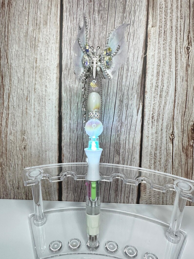 Puede incluir: Un bol&iacute;grafo decorativo con dise&ntilde;o de mariposa. El bol&iacute;grafo presenta una mariposa plateada con detalles transparentes e iridiscentes. El cuerpo del bol&iacute;grafo tiene un dise&ntilde;o blanco, transparente e iridiscente con una funci&oacute;n de iluminaci&oacute;n. El bol&iacute;grafo se exhibe en un soporte de pl&aacute;stico transparente.