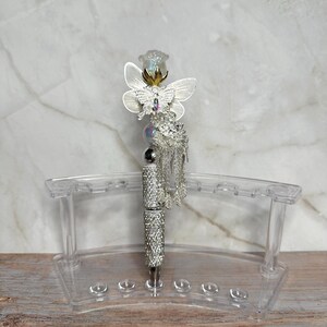 Op de afbeelding: Een decoratieve pen met een zilveren loop bezet met strass-steentjes. De pen is bekroond met een witte vlinder en een heldere, facetgeslepen bloem. Een zilveren kwast hangt aan de pen. De pen rust in een doorzichtige acryl pennenhouder.