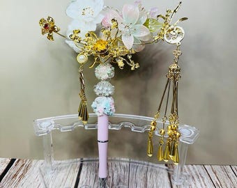 May include: A decorative pen with a light pink barrel, silver accents, and a floral design. The pen is displayed in a clear acrylic stand and adorned with gold-colored tassels and floral embellishments. The flowers are white, pink, and yellow.