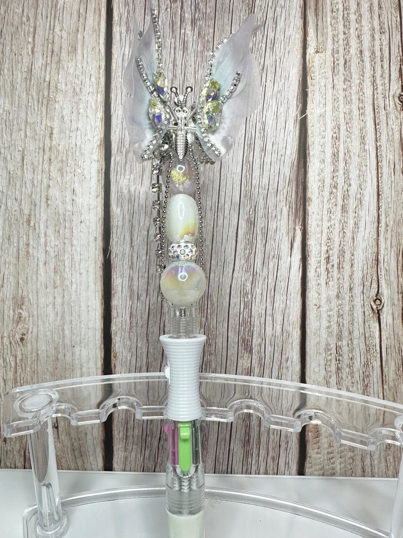Puede incluir: Un bol&iacute;grafo decorativo con dise&ntilde;o de mariposa. El bol&iacute;grafo presenta una mariposa plateada con pedrer&iacute;a transparente y amarilla y alas blancas y plumosas. El bol&iacute;grafo tiene un cuerpo blanco con una secci&oacute;n transparente.