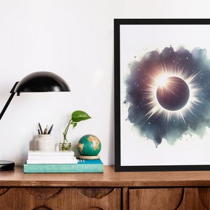 May include: A black framed print of a watercolour painting depicting a solar eclipse with a bright white halo and a dark circle in the centre. The background is a dark blue with white stars.
