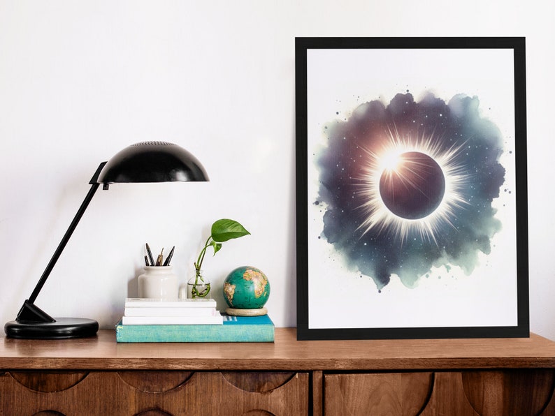 May include: A black framed print of a watercolour painting depicting a solar eclipse with a bright white halo and a dark circle in the centre. The background is a dark blue with white stars.