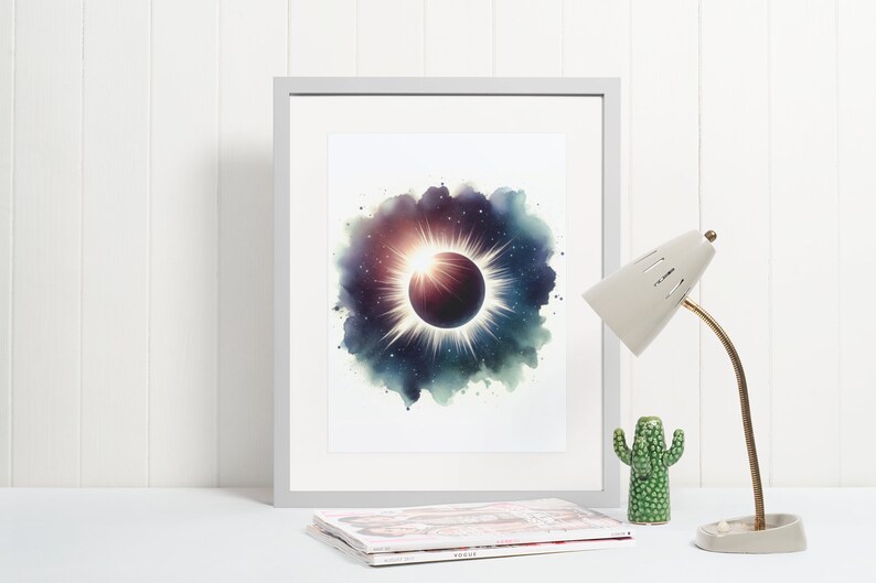 May include: A framed watercolour print of a solar eclipse with a dark blue, purple, and green background. The eclipse is surrounded by a white halo and stars. The print is in a white frame and is sitting on a white surface.