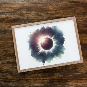 May include: A framed print of a watercolour painting depicting a solar eclipse. The eclipse is surrounded by a dark blue and green watercolour cloud with white stars. The sun is shining brightly behind the moon, creating a halo of light.