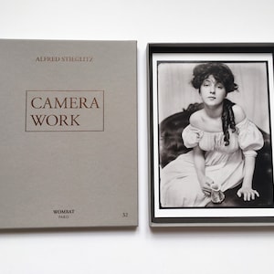 Op de afbeelding: Een grijze doos met de tekst "Alfred Stieglitz Camera Work" in bruin gedrukt op de voorkant. De doos is gelabeld met "Wombat Paris" en "32" in de rechterbenedenhoek. De doos bevat een zwart-witfoto van een vrouw die op een stoel zit en een kopje vasthoudt.