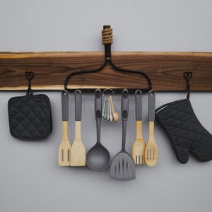 May include: A wooden wall-mounted rack with a black metal hanger and heart-shaped hooks. The rack is holding kitchen utensils, including wooden spoons, spatulas, and a ladle, as well as two black oven mitts.