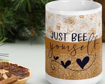 May include: White ceramic mug with a bee-themed design. The top section features a pattern of bees and honeycombs. The middle section is a gold color with the words "JUST BEE yourself" and two black hearts. The bottom section has a gold glitter effect.