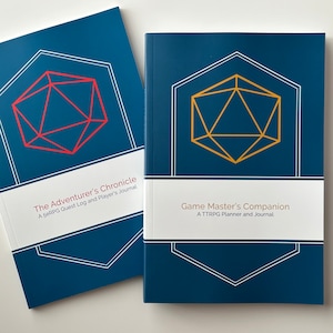 May include: Two blue hardcover journals with white and gold accents. The covers feature a stylized 20-sided die, a common element in tabletop role-playing games. The text on the covers reads "The Adventurer's Chronicle: A 5eRPG Quest Log and Player's Journal" and "Game Master's Companion: A TTRPG Planner and Journal".