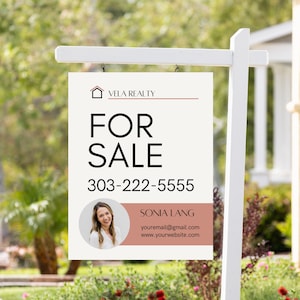 May include: A white real estate sign with black text that reads "Vela Realty", "For Sale", "303-222-5555", "Sonia Lang", "youremail@gmail.com", and "www.yourwebsite.com". The sign also features a photo of a woman.