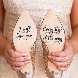 May include: A pair of champagne-colored high heels with rhinestone embellishments. The soles of the shoes have the phrases "I will love you" and "Every step of the way" written in elegant script. The shoes are held by a person wearing a wedding dress.