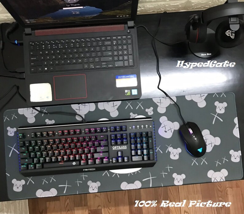 Op de afbeelding: Een zwart en grijs toetsenbord met regenboogverlichting en een zwarte muis op een grijze muismat met een herhalend patroon van witte teddyberen met een X op hun gezicht. De muismat heeft de tekst "HypedGate" in wit op een zwarte achtergrond. De tekst "100% Real Picture" staat in wit op een zwarte achtergrond onderaan de afbeelding.