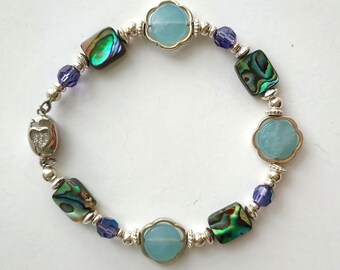 May include: A beaded bracelet featuring a mix of elements. The bracelet includes iridescent abalone shell pieces, light blue flower-shaped beads, and small purple beads. The bracelet is strung on a silver-colored chain.