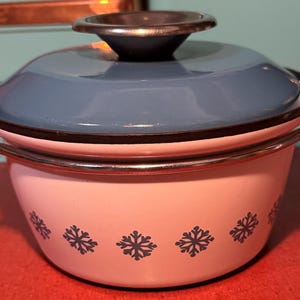 Puede incluir: Una olla vintage de esmalte blanco y azul con tapa. La olla tiene una tapa azul claro y un cuerpo blanco decorado con diseños de copos de nieve azul oscuro. Cuenta con asas metálicas.