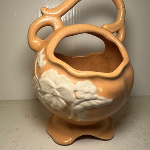 Könnte beinhalten: Pfirsichfarbener Keramikkorb mit großem Henkel und floralem Design. Der Korb hat einen Sockel und eine strukturierte, rissige Oberfläche. Das weiße Blumendesign befindet sich auf der Vorderseite des Korbes.