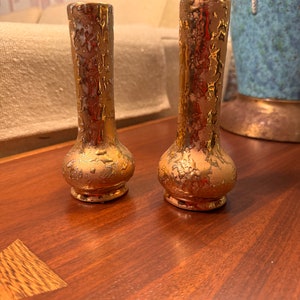 May include: Two small, gold-colored vases with a textured, dripped design. The vases have a bulbous base and a narrow neck. They are displayed on a dark wood surface, with a neutral-toned sofa and decorative vase in the background.