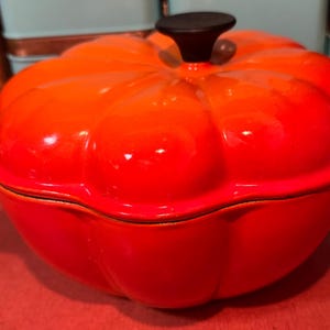 Può includere: Una casseruola olandese a forma di zucca, arancione e rossa lucida, con un pomello nero sul coperchio. La pentola ha una forma arrotondata e un manico laterale. La pentola è progettata per cucinare e servire.