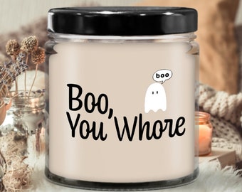 May include: A glass jar candle with a black lid. The candle has a light beige wax and features the text "Boo, You Whore" in black, along with a cartoon ghost saying "boo" in a speech bubble. The candle is a Halloween-themed item.
