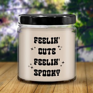 May include: A clear glass jar candle with a black lid. The candle has the words "FEELIN' CUTE FEELIN' SPOOKY" in black, stylized text with star accents. The candle sits on a wooden surface with a blurred green background.