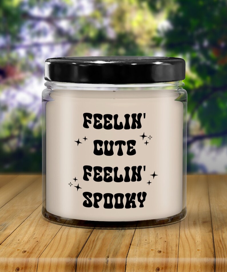 May include: A clear glass jar candle with a black lid. The candle has the words "FEELIN' CUTE FEELIN' SPOOKY" in black, stylized text with star accents. The candle sits on a wooden surface with a blurred green background.