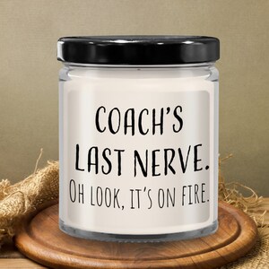 May include: A white candle in a glass jar with a black lid. The candle has a label that reads "Coach's Last Nerve. Oh look, it's on fire."