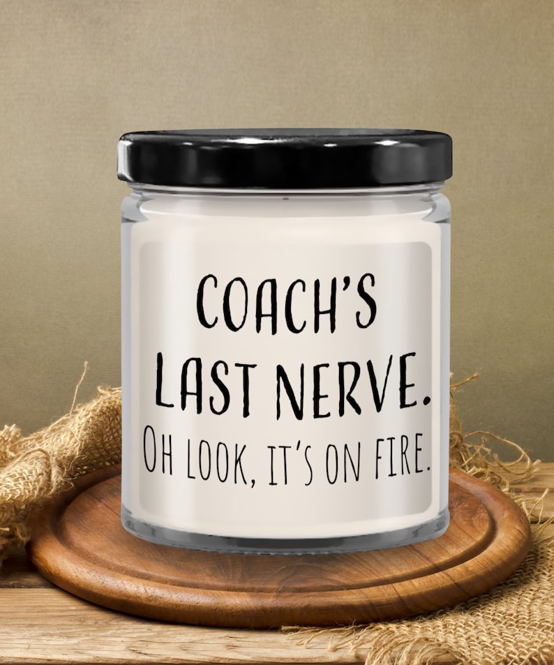May include: A white candle in a glass jar with a black lid. The candle has a label that reads "Coach's Last Nerve. Oh look, it's on fire."