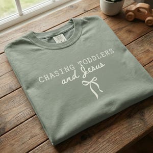 Può includere: T-shirt verde salvia con la scritta bianca "CHASING TODDLERS and Jesus" e un fiocco bianco. La maglietta è piegata su una superficie di legno. È del marchio Comfort Colors.