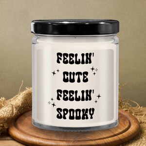 May include: A clear glass jar candle with a black lid, sitting on a wooden coaster. The candle has a white wax interior and features the text "FEELIN' CUTE FEELIN' SPOOKY" in black, with star accents. The background includes burlap and a wooden surface.