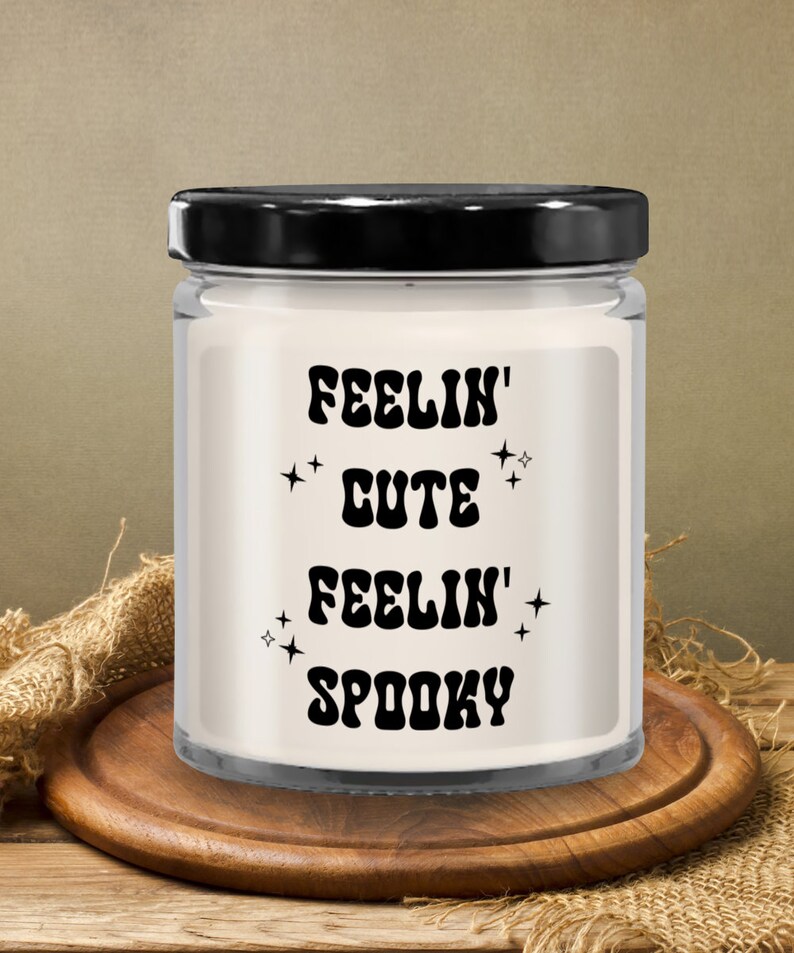 May include: A clear glass jar candle with a black lid, sitting on a wooden coaster. The candle has a white wax interior and features the text "FEELIN' CUTE FEELIN' SPOOKY" in black, with star accents. The background includes burlap and a wooden surface.