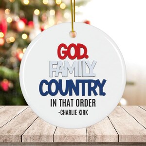 Puede incluir: Un adorno navideño blanco y redondo con las palabras "GOD, FAMILY, COUNTRY" en rojo, gris y azul, con la frase "IN THAT ORDER" y el nombre "-CHARLIE KIRK" debajo. El adorno cuelga de un cordón dorado.