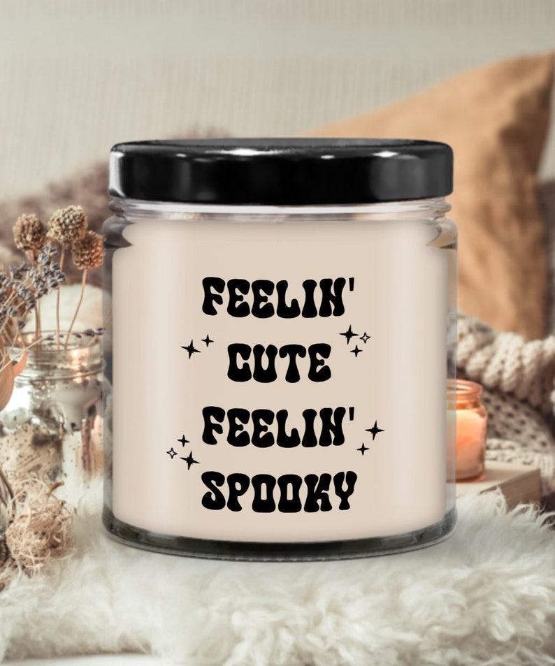 May include: A glass jar candle with a black lid. The candle has a light pink wax and features the text "FEELIN' CUTE FEELIN' SPOOKY" in black, retro-style lettering, with star accents. The candle is set against a soft, neutral background.