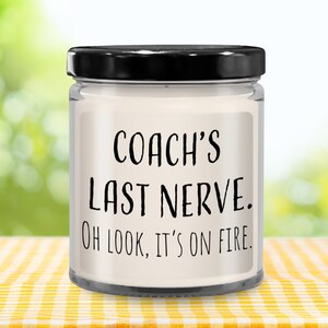 May include: A white candle in a clear glass jar with a black lid. The candle has a label that reads "COACH'S LAST NERVE. OH LOOK, IT'S ON FIRE."