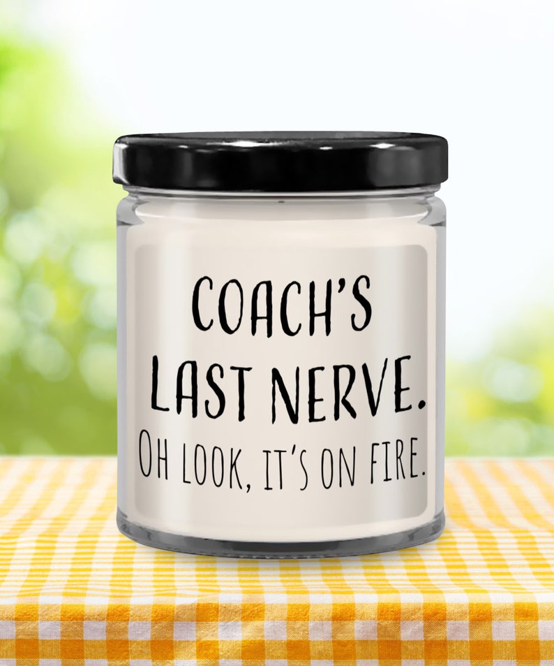 May include: A white candle in a clear glass jar with a black lid. The candle has a label that reads "COACH'S LAST NERVE. OH LOOK, IT'S ON FIRE."