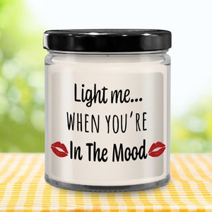 May include: A white candle in a glass jar with a black lid. The candle has a label that says "Light me... When you're In The Mood" with red lips on either side of the text.