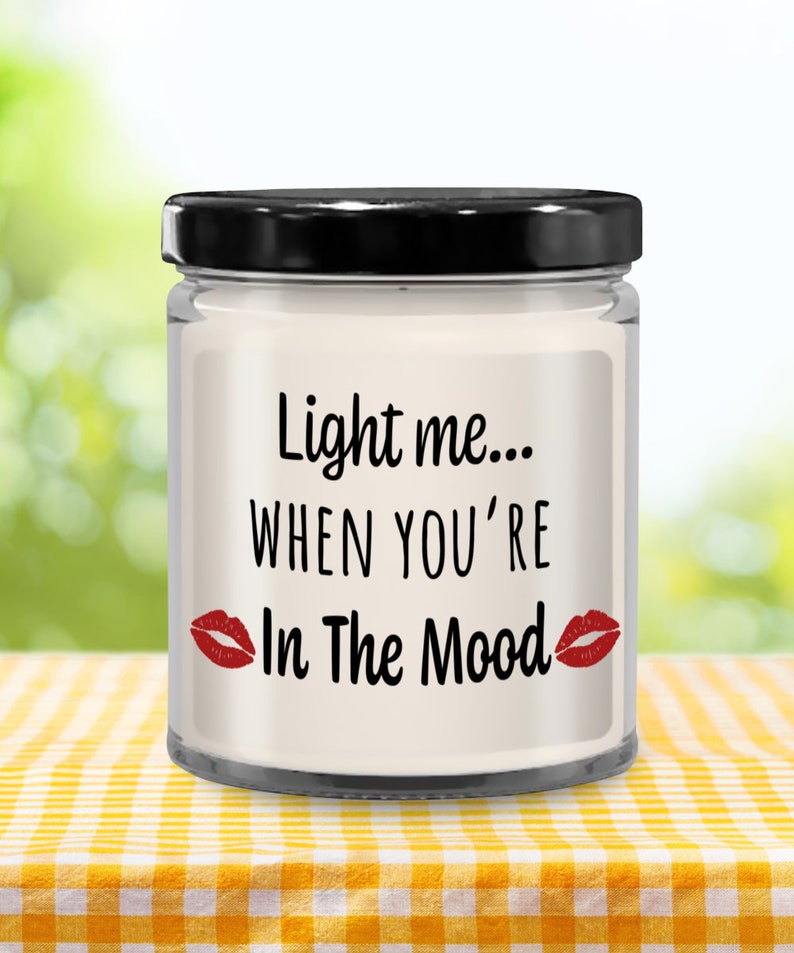 May include: A white candle in a glass jar with a black lid. The candle has a label that says "Light me... When you're In The Mood" with red lips on either side of the text.