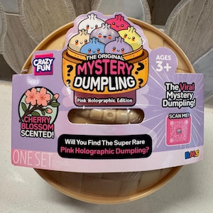 以下が含まれることがあります： The Original Mystery Dumpling Pink Holographic Editionが入った丸いベージュ色の容器。紫色のパッケージには、餃子のキャラクター、桜の香りのラベル、QRコード、そして「Will You Find The Super Rare Pink Holographic Dumpling?」というテキストが記載されています。このおもちゃは3歳以上を対象としています。