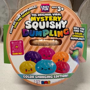 以下が含まれることがあります： Mystery Squishy Dumplingというおもちゃの丸いオレンジ色のパッケージ。前面には「The Original Viral Mystery Squishy Dumpling」、「Color Changing Edition!」、「Ages 3+」などのテキストが記載されています。カラフルな餃子型のスクイーズおもちゃが描かれています。