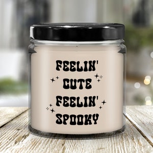 May include: A glass jar candle with a black lid. The candle has a cream-colored wax and features the text "FEELIN' CUTE FEELIN' SPOOKY" in black, retro-style lettering, with star accents. The candle sits on a light-colored wooden surface.