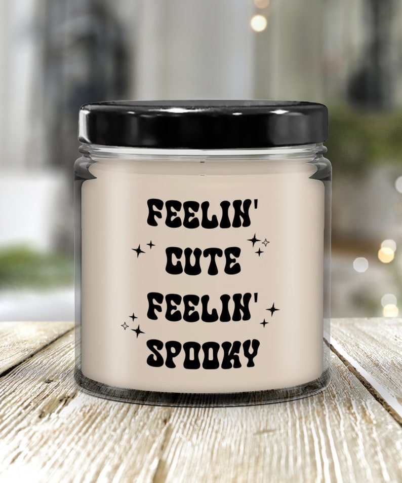 May include: A glass jar candle with a black lid. The candle has a cream-colored wax and features the text "FEELIN' CUTE FEELIN' SPOOKY" in black, retro-style lettering, with star accents. The candle sits on a light-colored wooden surface.