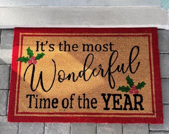 May include: A red and brown doormat with the text "It's the most wonderful time of the year" in black lettering. The doormat has a green holly design with red berries on each side of the text.