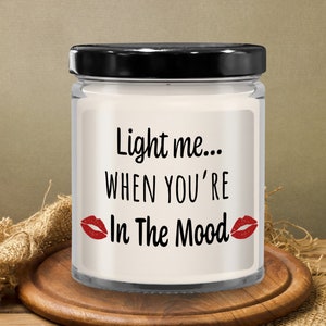 May include: A white candle in a glass jar with a black lid. The candle has the text "Light me... When you're In The Mood" with red lips on either side of the text.